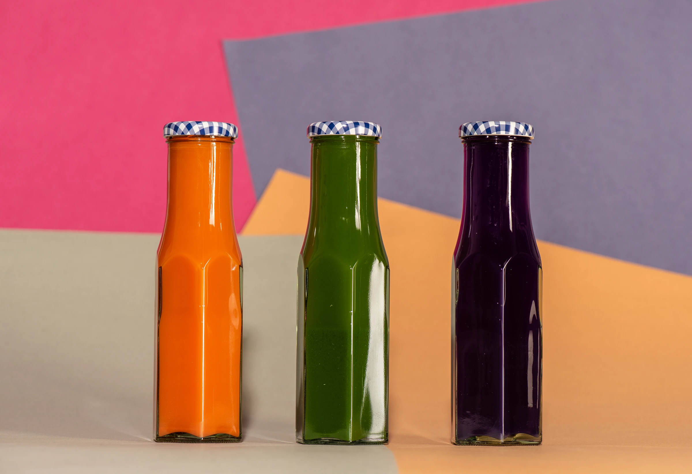 Three glass bottles containing orange, green and purple liquids against coloured paper